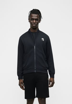 Giovane uomo con dreadlocks che indossa una giacca nera con cerniera e un piccolo logo bianco, e pantaloncini neri, in piedi contro uno sfondo semplice.