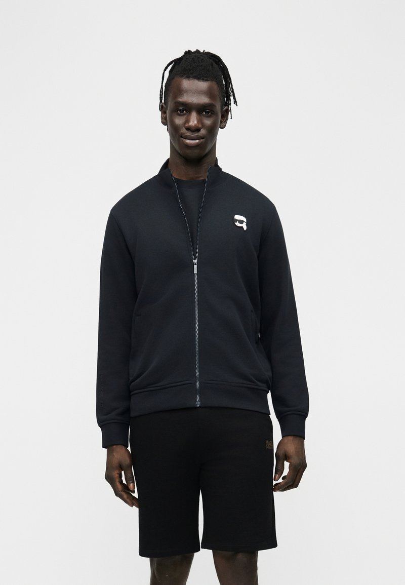 Νέος άνδρας με dreadlocks που φοράει μαύρο τζάκετ με φερμουάρ και μικρό λευκό λογότυπο, καθώς και μαύρο σορτς, στέκεται μπροστά σε ένα απλό φόντο.