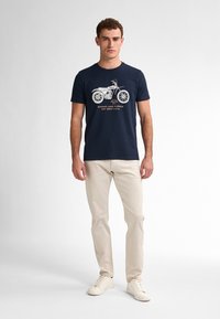 Tmavě modré bavlněné tričko s bílou grafikou motocyklu a textem. V kombinaci se světlými béžovými kalhotami a bílými teniskami.