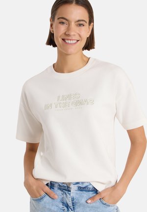 Femme souriante aux cheveux courts et bruns porte un t-shirt blanc avec le texte "LINES IN THE SANDS" et un jean bleu clair, les mains dans les poches.