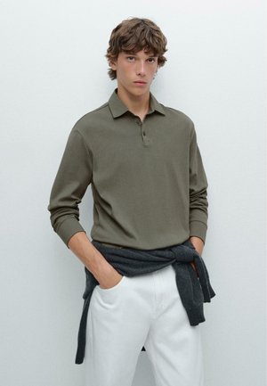 Olive green long-sleeve polo shirt with a collar, showcasing a soft texture. White trousers are worn with a dark gray sweater tied at the waist.
