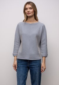 Gestreept pullover-topje met een boothals, voorzien van afwisselende blauw-witte zigzagpatronen en driekwartmouwen. Gecombineerd met een blauwe spijkerbroek.