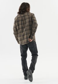 Geruite knoopshirt in olijfgroen en bruin, gecombineerd met zwarte jeans en zwarte laarzen. Het shirt heeft een relaxte pasvorm en een achterpand.