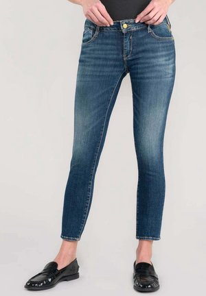 Personne portant un jean bleu slim et des mocassins noirs, les mains ajustant la taille, debout sur un fond clair.