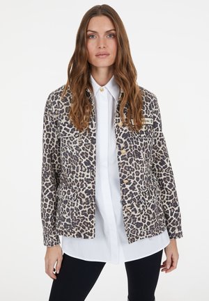 Frau mit langen braunen Haaren, die eine Jacke mit Leopardenmuster über einem weißen Hemd und schwarzen Hosen trägt, vor einem einfarbigen Hintergrund stehend.