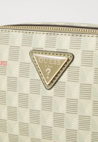 Geanta Guess U.S.A. cu un fundal crem, model cu herringbone, logo triunghiular aurit și o textură rafinată, cu cusături intricate.