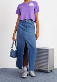 Purple cropped t-shirt with logo, paired with a long, light blue denim skirt featuring a front slit, and gray athletic shoes. Small denim bag.