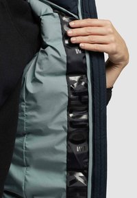 Blauwe jas met een lichtgroene binnenkant. De voering heeft een zwarte stoffen strook met witte branding, wat een textuurcontrast geeft.