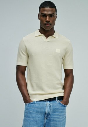 Homme portant un polo en maille à manches courtes de couleur crème, légèrement rentré dans un jean bleu clair, les mains dans les poches, sur un fond uni.