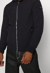 Sort hættejakke med lynlås foran, udstyret med sidelommer og ribbede manchetter. Fremstillet af et glat, letvægtsstof.