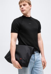 Νέος άνδρας με μαύρο πουκάμισο κοντών μανικιών κρατάει μια μαύρη τσάντα χειρός με λογότυπο Hugo, φορά ανοιχτόχρωμο μπλε τζιν και κοιτά προς τα αριστερά.