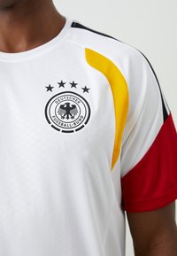 Λευκή αθλητική φανέλα με κίτρινες και κόκκινες λεπτομέρειες. Φέρει ένα μαύρο λογότυπο με έναν αετό και πέντε αστέρια. Κατασκευασμένη από υλικό με υφή που αναπνέει.