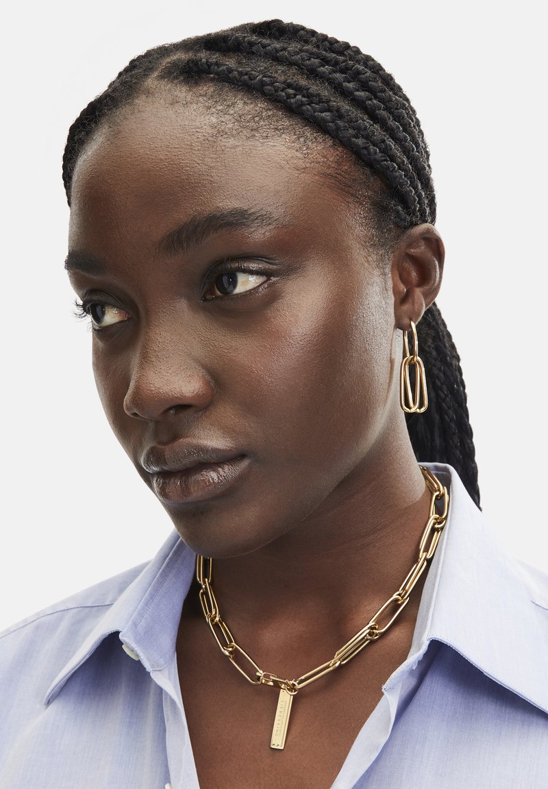 Collier en chaîne dorée avec des maillons rectangulaires et un pendentif. Porté sur une chemise à col bleu clair, avec des cheveux tressés complexes.