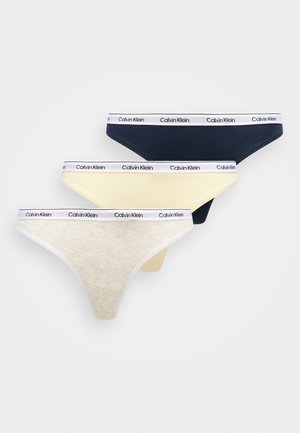 Bralette en coton gris avec fines bretelles et bande logo, associée à un string jaune. Le mannequin se tient pieds nus sur un tapis gris à côté d'une chaise douce et incurvée.