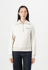 Light beige sweatshirt with a zip collar, ribbed cuffs, and hem. Features an embroidered logo on the chest. Paired with dark jeans.