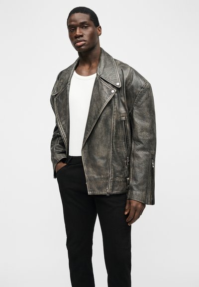 Jeune homme portant une veste en cuir grise délavée surdimensionnée, un t-shirt blanc et un pantalon noir, debout avec une main dans la poche.