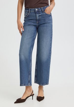 Femme portant un jean cropped bleu à jambes larges avec un haut marron et des talons slingback pointus marron, debout devant un fond uni.