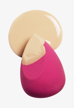 Éponge de maquillage en forme de goutte, rose d'un côté et beige de l'autre. Une goutte circulaire de fond de teint beige se trouve à proximité.