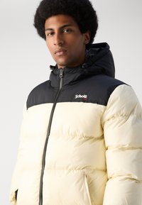 Giacca imbottita con cappuccio nero e parte superiore nera, corpo color crema e chiusura con zip. Presenta il logo sul petto e una texture trapuntata.