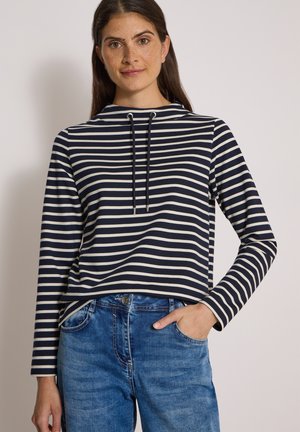 Femme aux longs cheveux bruns portant un haut à rayures bleu marine et blanc à manches longues et un jean bleu, une main dans la poche.