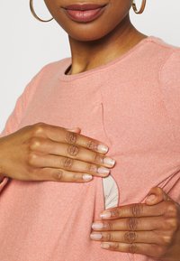 Světlounce růžový svetr s texturovaným povrchem. Nenápadný boční otvor odhaluje béžovou vrstvu pod ním. Ruce umístěné na otvoru.