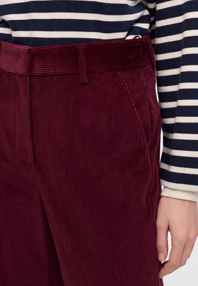 Gros plan d'une personne portant un pantalon en velours côtelé bordeaux et une chemise à manches longues rayée bleu marine et blanc, la main détendue le long du corps.
