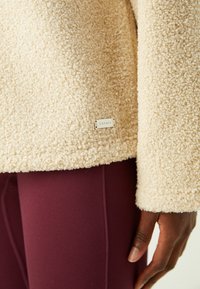 Pullover in pile beige con superficie testurizzata, dotato di un piccolo logo in argento sul bordo. Abbinato a leggings borgogna.