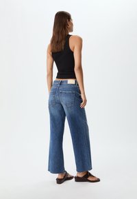 Vida byxor i blå denim med vida ben och rå fåll, med hög midja och detaljer i form av lapplådor. Stylen är kompletterad med en svart ärmlös topp och bruna sandaler.