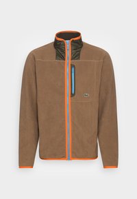 Brun fleecjacka med hög krage, blå dragkedja och orange detaljer. Har en grön bröstficka och strukturerad tyg.