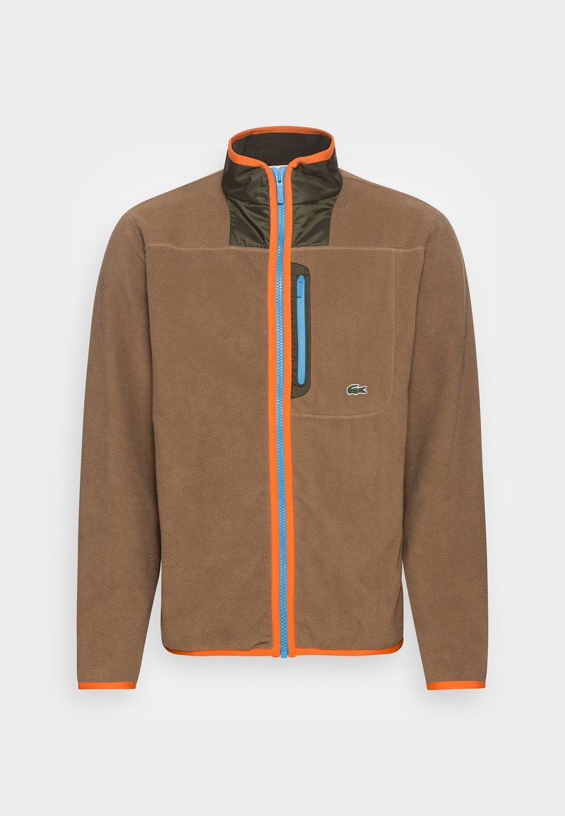 Brun fleecjacka med hög krage, blå dragkedja och orange detaljer. Har en grön bröstficka och strukturerad tyg.