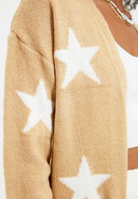 Beige stickad cardigan med vitt stjärnmönster, som har en texturerad yta och lös passform. Inkluderar en kraftig nacke och mjukt tyg.