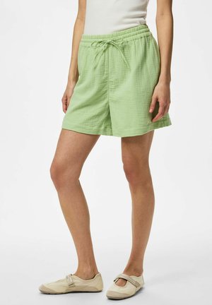 Kvinne iført lysegrønne teksturerte shorts med snøring og beige Mary Jane-stil sko, står mot en hvit bakgrunn.
