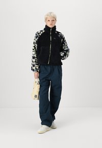 Chaqueta de forro polar con diseño en bloques de color en navy y blanco, con cuello de cremallera, combinada con pantalones azul oscuro y llevando un bolso de mano crema.