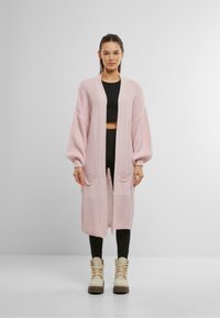 Gebreide cardigan in zacht roze met lange mouwen, een open voorkant en twee voorzakken. Geweven ontwerp; gecombineerd met zwarte leggings en beige laarzen.