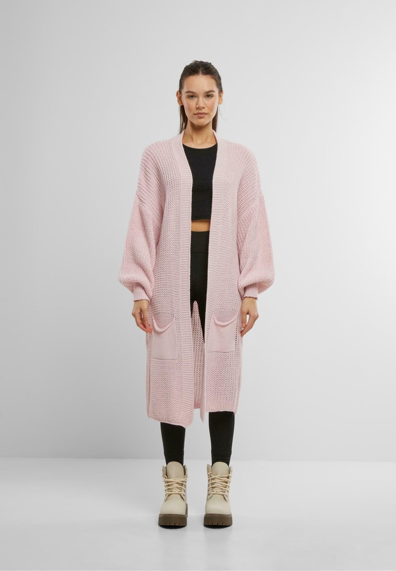 Gebreide cardigan in zacht roze met lange mouwen, een open voorkant en twee voorzakken. Geweven ontwerp; gecombineerd met zwarte leggings en beige laarzen.