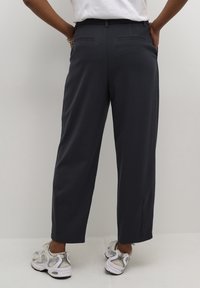 Pantalon large gris foncé en tissu lisse, avec des poches arrière et une coupe droite. Associé à des baskets blanches et argentées.