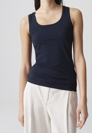 Femme portant un débardeur bleu marine ajusté sans manches et un pantalon blanc ample sur un fond gris clair uni.