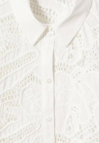 White shirt featuring intricate lace detailing, a classic collar, and button closure. Made of lightweight fabric with a textured finish.
