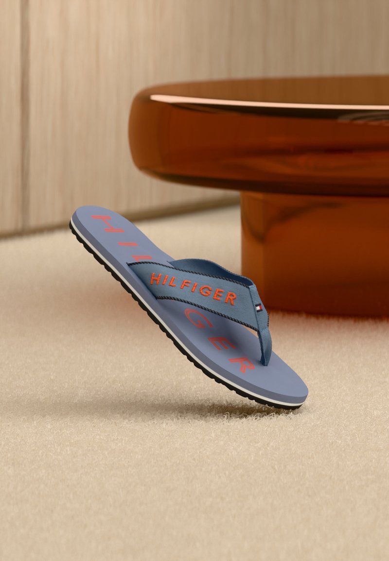 Tongs bleues avec le texte orange "HILFIGER" et le logo sur la bride, posées sur un tapis beige à côté d'une table ronde marron.
