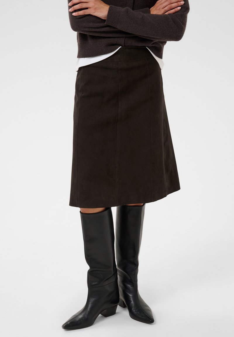 Femme portant une jupe marron foncé jusqu'aux genoux, des bottes noires montantes et un pull sombre, les bras croisés.