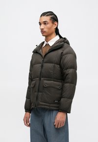 Mørkebrun polstret jakke med glidelås foran, justerbar hette og to knappede brystlommer. Mykt materiale med quiltet tekstur.