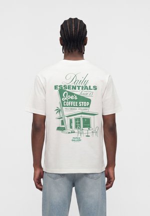 Mand med flettet hår iført hvid t-shirt med grønt motiv af Joe’s Coffee Stop-skilt og bygning, kombineret med lyseblå jeans.