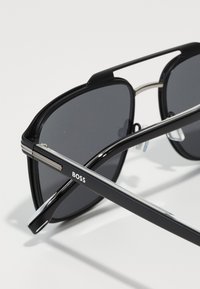 Ochelari de soare negri cu un design elegant și unghiular. Lentile gri și un cadru accentuat cu argint. Logo-ul "BOSS" pe braț.