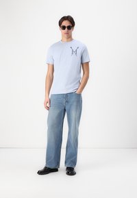 Šviesiai mėlynas medvilninis marškinėliai su krūtinės piešiniu, derinami su laisvais mėlynais džinsais ir juodais odiniais batais. Modelis dėvi saulės akinius.