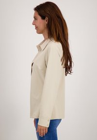Lichtbeige langemouwen shirt met een kraag, zijknopen en een relaxte pasvorm. Ggedragen met blauwe jeans, die de soepele stofstructuur tonen.