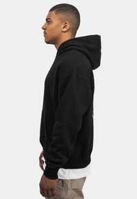 Sudadera negra con una capucha prominente, puños acanalados y un bolsillo frontal; presenta una ligera textura y un diseño minimalista.