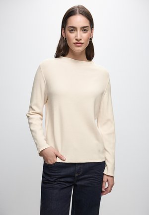 Hellbeige, geripptes Sweatshirt mit langen Ärmeln, Rundhalsausschnitt und lockerer Passform, kombiniert mit dunkelblauen Jeans.