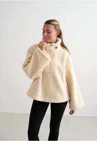 Krémový fleecový pulovr s vysokým límcem, polo-zip uzávěrem a bočními kapsami. Disponuje širokými rukávy a texturovaným, plyšovým povrchem.