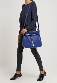 Modrá kožená kabelka s nastavitelným popruhem, hladkým povrchem, stříbrnými detaily a zipovým uzávěrem. Zobrazena na neutrálním pozadí.