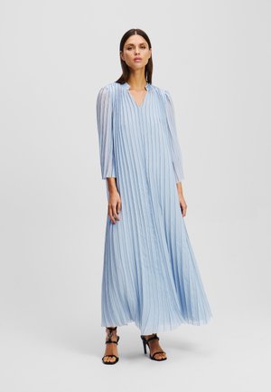 Robe maxi plissée bleu clair avec un décolleté en V, des manches longues et de subtiles rayures verticales. Associée à des sandales à talons noirs.